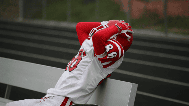 stack.com – sports tryouts mental toughness young football athlete showing frustration by grabbing helmet while sitting on bench